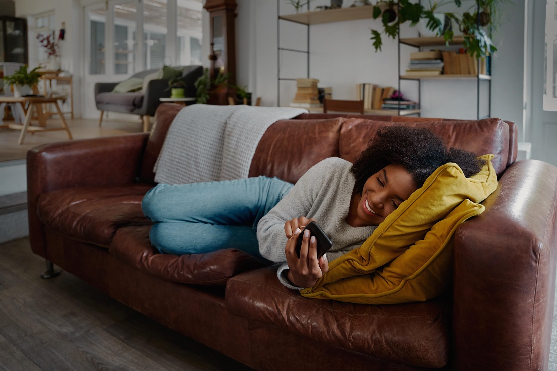 A person smiles while reclining on a couch looking at a mobile phone